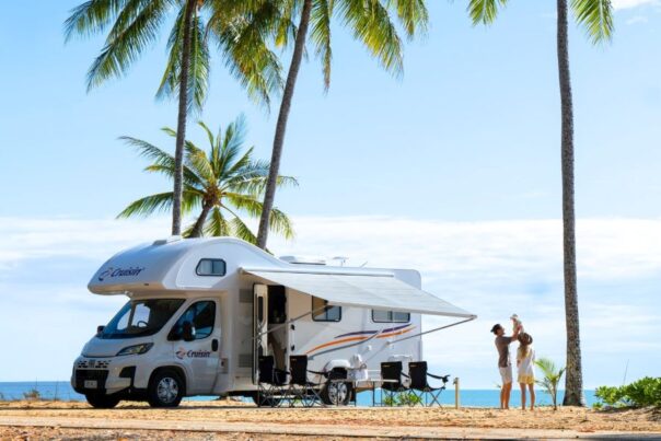 Vader tilt jong kind op naast Cruisin' 6 Berth Deluxe camper die tussen palmbomen aan het strand geparkeerd staat