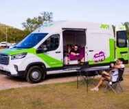 Jucy Coaster staat op een camping met de zij- en achterdeuren open, een man ligt op het bed en een man zit op een campingstoel buiten