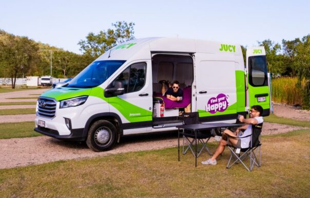 Jucy Coaster staat op een camping met de zij- en achterdeuren open, een man ligt op het bed en een man zit op een campingstoel buiten
