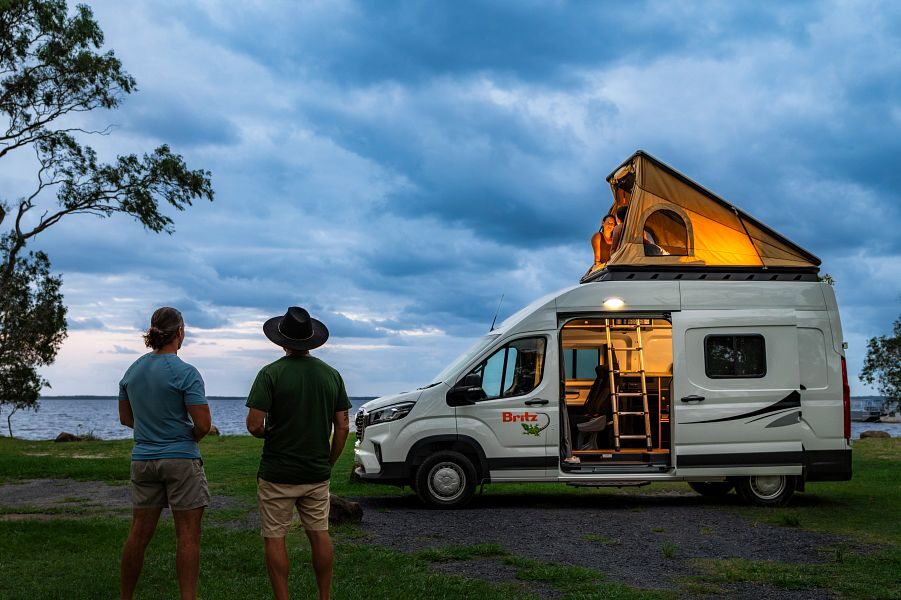 Britz Voyager staat geparkeerd in schemer met daktent omhoog en licht aan binnen, twee mannen lopen op de camper af