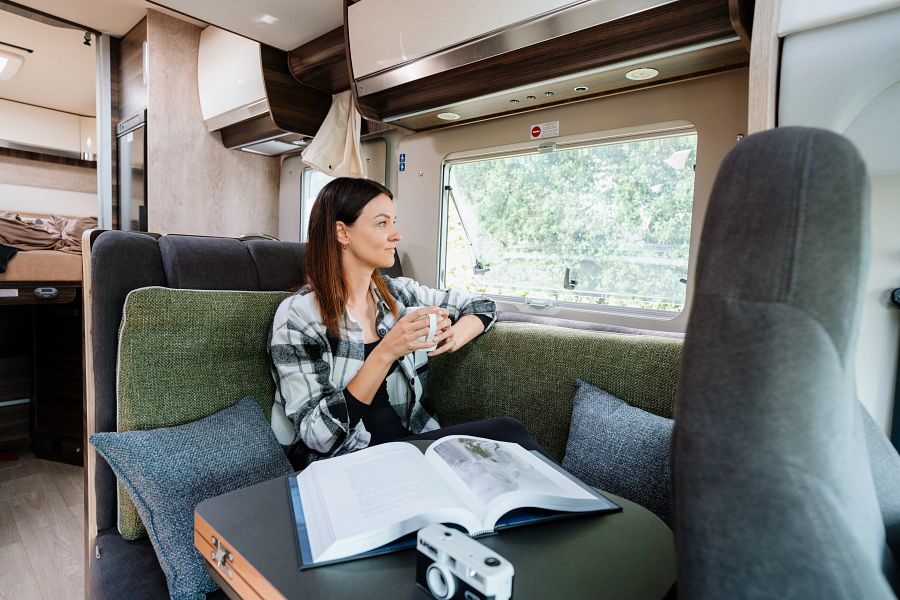Een vrouw zit in de zithoek van de Britz Wanderer camper met een boek op tafel en een mok in haar hand kijkend door het raam naar buiten