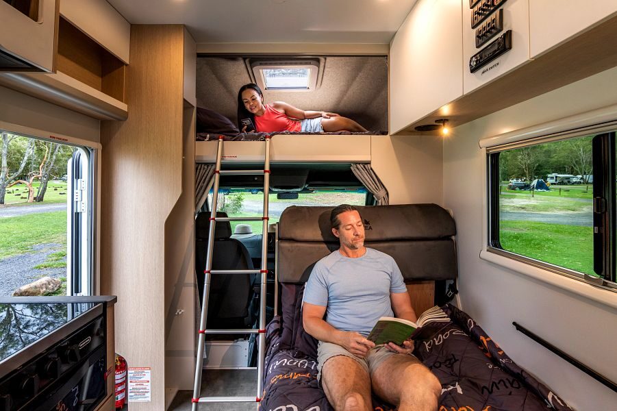 Een vrouw ligt op het tweepersoonsbed in de alkoof van de Maui River camper die bereikbaar is met een ladder en een man ligt een boek te lezen op het bed in het midden van de camper