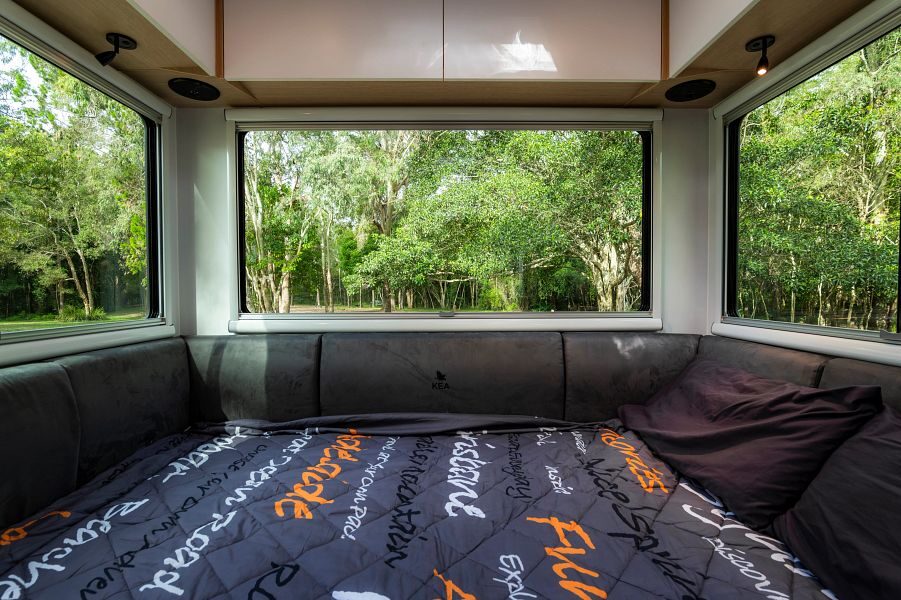Het opgemaakte tweepersoonsbed achterin de Maui River camper met grote ramen met uitzicht op de groene natuur