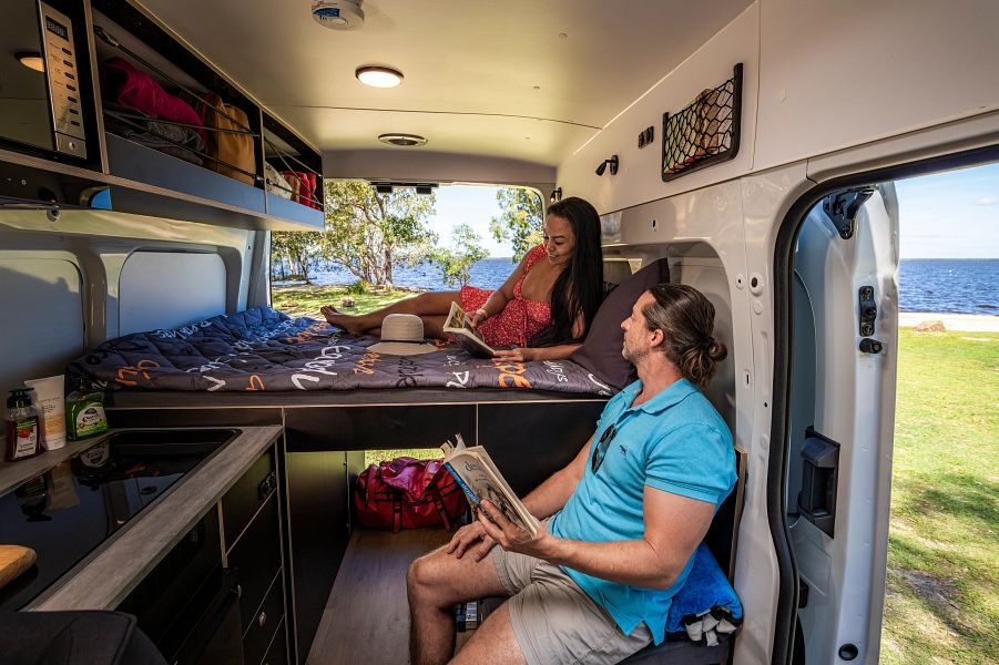 Een vrouw ligt op het tweepersoonsbed van de Apollo Hitop camper in Australië een boek te lezen terwijl een man zit op het afgesloten toilet blok