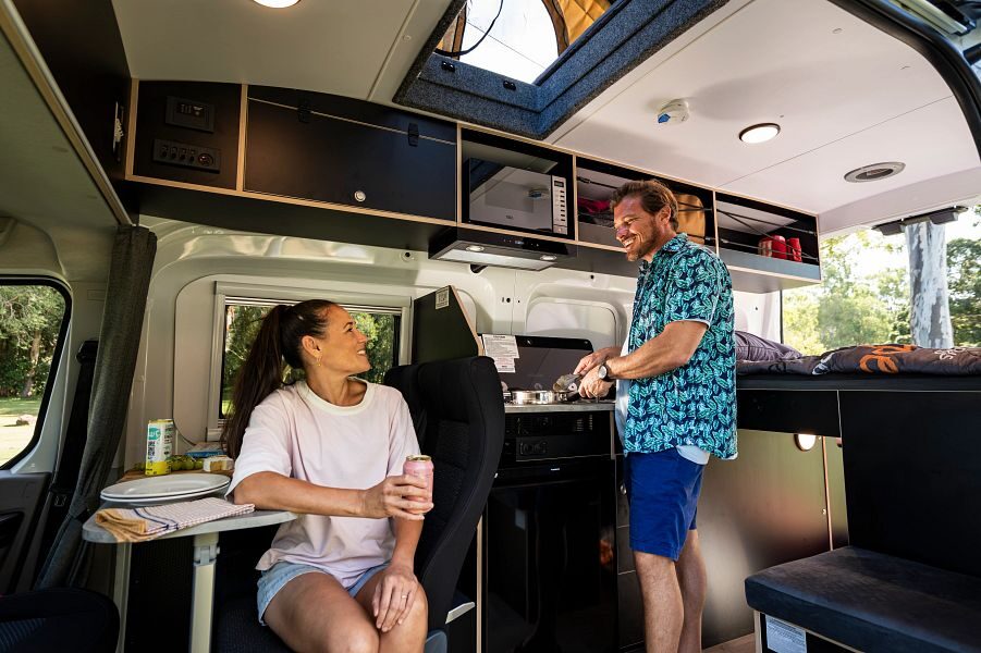 Een man staat in de keuken van de Mighty Double Down camper te koken terwijl een vrouw in de zithoek aan tafel zit met een blikje drinken in haar hand