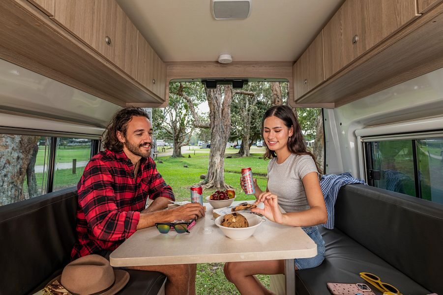 Een vrouw pakt een koekje terwijl ze samen met haar man aan tafel op de banken zit achterin de Mighty Duo camper met de achterdeuren open