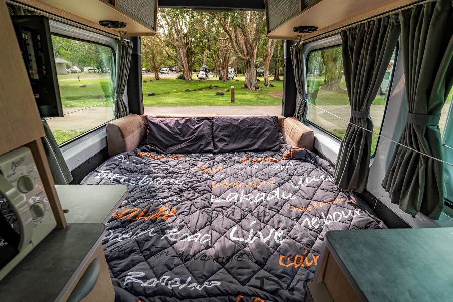 Een opgemaakt tweepersoonsbed aan de achterkant van de Mighty Deuce Plus camper met de achterdeuren open op de camping