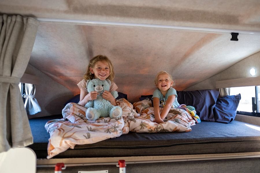 Twee kinderen zitten met een knuffel op het tweepersoonsbed in de alkoof boven de cabine van de Mighty Double Up camper