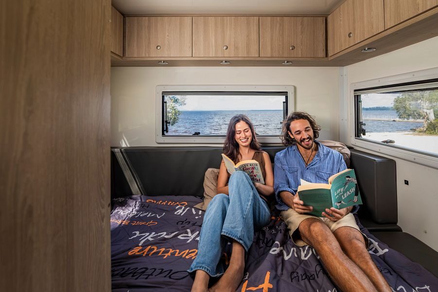 Een man en vrouw lezen beide een boek terwijl ze op het tweepersoonsbed achterin de Mighty Double Up camper zitten