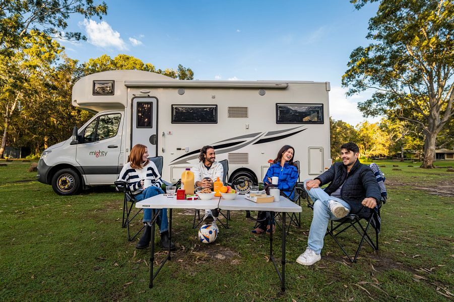 Een groep vrienden zit buiten aan tafel in de campingstoelen voor de Mighty Big Six camper die geparkeerd staat op het gras