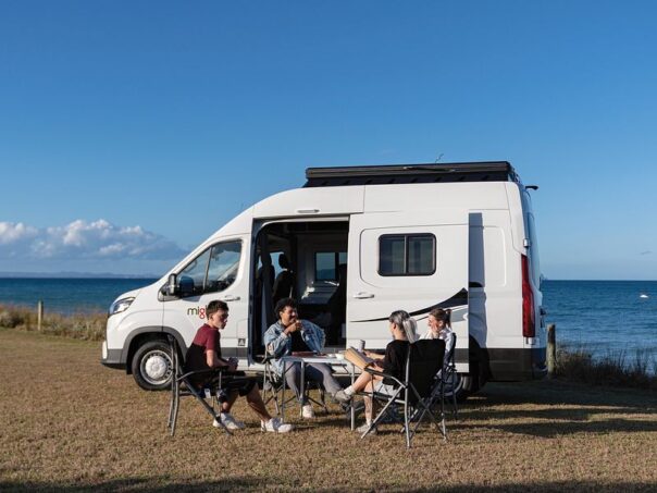 Een vriendengroep zit voor de Mighty Lite DeLITE camper met uitzicht op de zee in Nieuw-Zeeland