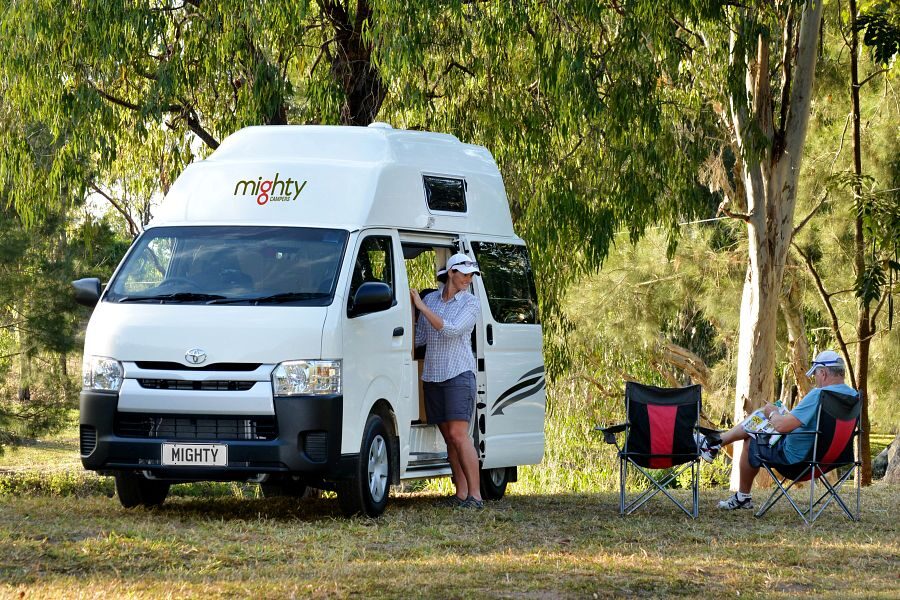 Een vrouw staat bij de open zijdeur van de Mighty Lite DeLITE camper in Australië terwijl haar man in een campingstoel zit