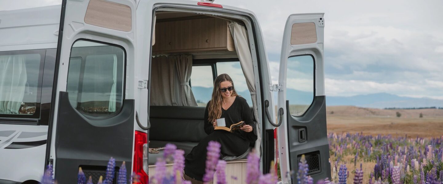 Vrouw zit in Britz Venturer camper met deur open en kijkt naar de lupines