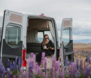 Vrouw zit in Britz Venturer camper met deur open en kijkt naar de lupines