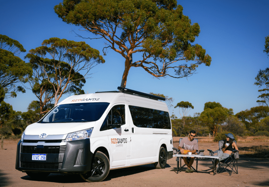 De camper is geparkeerd voor een tussenstop in een omgeving met bomen en twee personen zitten op hun campingstoelen en nuttigen een beker jus d'orange