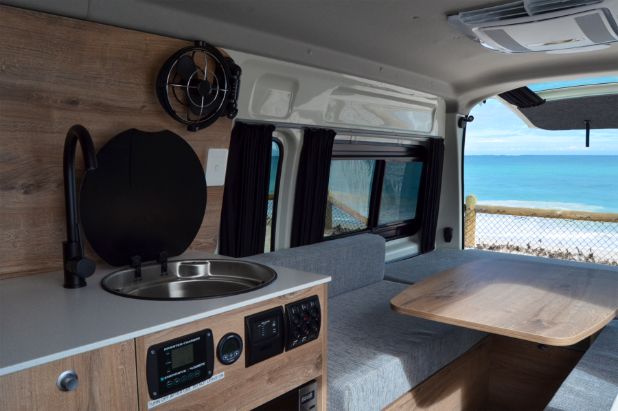 Het interieur van de achterzijde van de camper met onder andere een bedieningspaneel, een kraan met wasbak, een ventilator en airconditioning voor verkoeling en een zitgedeelte met tafel met de achterklep geopend