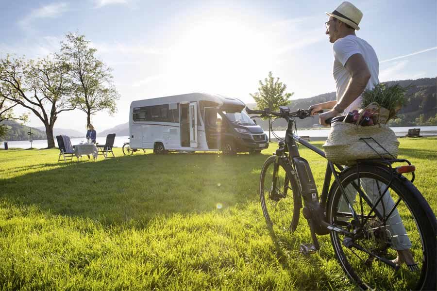 Een man loopt met zijn fiets richting zijn vrouw bij de McRent Comfort Plus camper die geparkeerd staat op het gras naast een meer