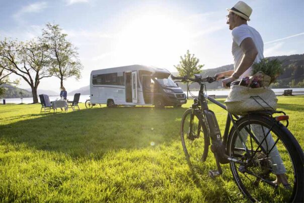 Een man loopt met zijn fiets richting zijn vrouw bij de McRent Comfort Plus camper die geparkeerd staat op het gras naast een meer