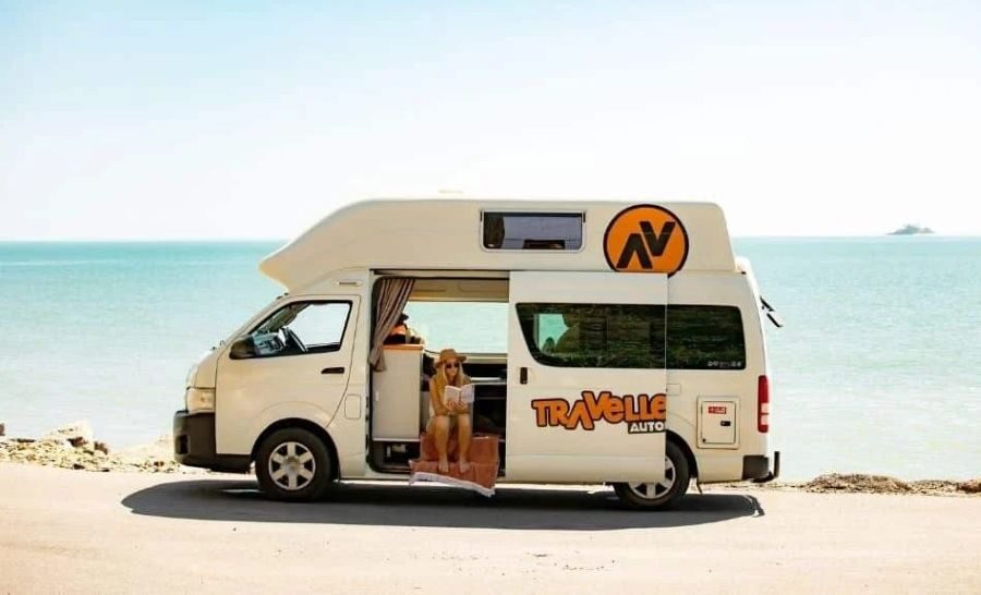 Vrouw leest een boek in de open zijdeur van de Travellers Autobarn Hi5 die geparkeerd staat aan het water