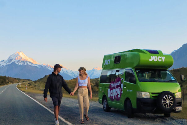 Een koppel loopt hand in hand naast een weg en de geparkeerde Jucy Condo camper in Nieuw-Zeeland