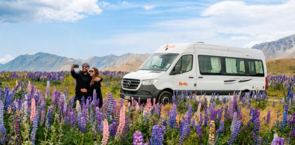 Stel maakt een selfie voor een Britz camper met lupines op de voorgrond