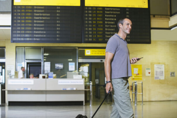 Man trekt koffer op luchthaven voor het vertrekbord en met telefoon in de hand